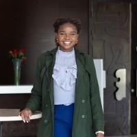 Louisa Konchellah posing facing the camera wearing a hunter green jacket over blue pants and a periwinkle blouse.